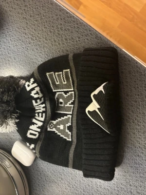 Svart mössa med tofs och Åre-tryck - Säljer en svart stickad mössa från OneWear med vit och grå text 'ÅRE' och en stor tofs på toppen. Mössan har ribbad kant och ett broderat bergmotiv på sidan. Perfekt för kalla dagar i fjällen eller stan.