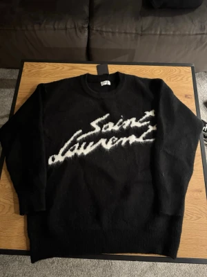 Svart Saint Laurent tröja - Svart stickad tröja från Saint Laurent med vit logotyp i graffiti-stil över bröstet. Tröjan har rund halsringning och ribbade muddar vid ärmslut och nederkant. Perfekt för dig som gillar exklusiva streetwear-vibbar. 