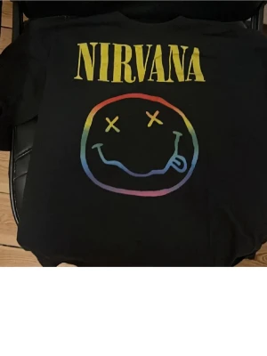 Svart Nirvana t-shirt med regnbågstryck - Cool svart t-shirt med Nirvana-tryck i gult och regnbågsfärgad smiley på bröstet. Klassisk passform och rund hals. Perfekt för dig som gillar bandmerch och vill sticka ut med färg.