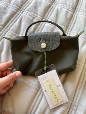 Grön väska från Longchamp - Snygg grön väska från Longchamp i nylon med detaljer i läder. Väskan har ett lock med tryckknapp och ett litet handtag upptill. Klassisk och stilren design med Longchamp-logga på locket. Perfekt för att hålla koll på kort och småsaker.