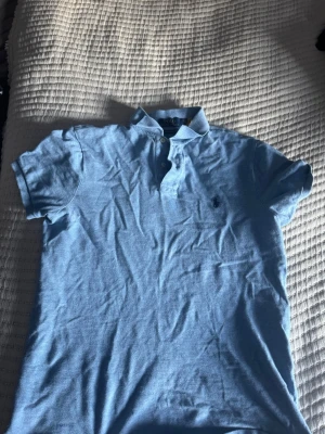 Ljusblå pikétröja från Ralph Lauren - Klassisk ljusblå pikétröja från Ralph Lauren med korta ärmar och krage. Tröjan har knappar vid halsen och den ikoniska broderade loggan på bröstet. Perfekt till jeans eller shorts för en avslappnad men snygg look.