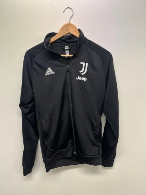 Svart Juventus träningsjacka Adidas - Snygg svart träningsjacka från Adidas med Juventus och Jeep-logga på bröstet. Jackan har hel dragkedja, ståkrage och vita detaljer på axlarna. Perfekt för fotbollsträning eller chill. Tillverkad i lätt och bekvämt material.
