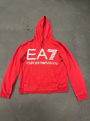 Röd EA7 Emporio Armani hoodie - Snygg röd hoodie från EA7 Emporio Armani med stor vit logga framtill och mindre logga bak. Klassisk huva med dragsko och känguruficka. Tillverkad i mjukt bomullsmaterial, perfekt för en avslappnad och trendig look.