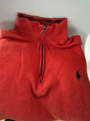 Röd stickad tröja från Polo Ralph Lauren - Snygg orange/röd stickad tröja från Polo Ralph Lauren med halv dragkedja och hög krage. Tröjan har det klassiska broderade logotypen i svart på bröstet och är tillverkad i bomull. Perfekt för lager-på-lager och chill stil.