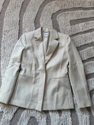 Krämvit glittrig kavaj från Kappahl - Stilren krämvit glittrig kavaj från Kappahl med klassisk passform och en knapp framtill. Kavajen har spetsiga slag, två fickor och är tillverkad i ett glansigt material som ger en elegant look. Perfekt att styla med både jeans och kjol. Små axelvaddar. Helt oanvänd. 