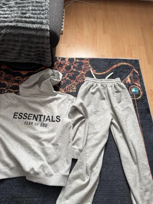 Grått Essentials Fear of God set - Mysigt grått loungewear-set från Essentials Fear of God. Setet består av en hoodie med tryck på bröstet och ryggen samt matchande mjukisbyxor med resår i midjan och fickor. Perfekt för chill dagar hemma. Materialet är mjukt och bekvämt bomullsblandat tyg.