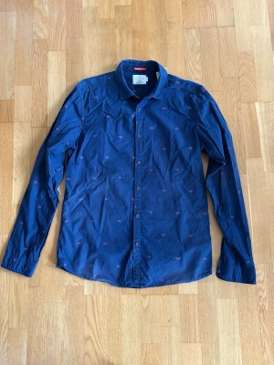 Blå skjorta från Scotch & Soda - Snygg marinblå skjorta från Scotch & Soda med små rosa broderade detaljer. Skjortan har klassisk krage, långa ärmar och knäppning framtill. Perfekt för dig som vill ha en stilren men ändå unik look.