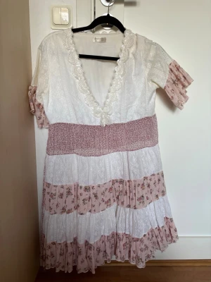  blommig klänning  - Söt klänning med v-ringning och spetskant, i vitt och ljusrosa med blommiga volanger. 