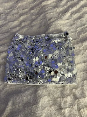 Silvrig paljettkjol med stora paljetter - Glittrig minikjol i silver med stora runda paljetter över hela tyget. Kjolen har en tight passform och är fodrad med vitt tyg på insidan för extra komfort. Perfekt för dig som vill sticka ut och glänsa på nästa fest. Aldrig använt pågrund av att de inte är min stil! 