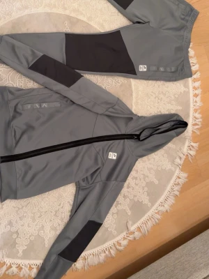 Grå träningsset från Nike med svarta detaljer - Snyggt grått träningsset från Nike med svarta detaljer på ärmar och ben. Setet består av en zip-hoodie och matchande byxor, båda med Nike-logga. Materialet är mjukt och stretchigt, perfekt för träning eller chill. Jackan har dragkedja och fickor framtill.