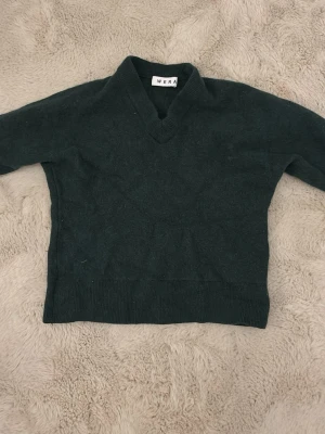 Mörkgrön stickad tröja från Wera - Mysig mörkgrön stickad tröja från Wera med v-ringad hals och långa ärmar. Tröjan har ribbade muddar vid ärmslut och nederkant, vilket ger en snygg och enkel look. Perfekt att matcha med jeans eller kjol för en chill stil.