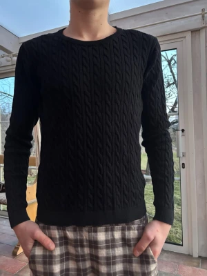 Svart kabelstickad tröja - Säljer en svart kabelstickad tröja med rund hals och långa ärmar. Tröjan har ett klassiskt flätmönster över hela plagget och ribbade muddar vid ärmslut och nederkant. Perfekt för dig som gillar stilrena och tidlösa plagg.