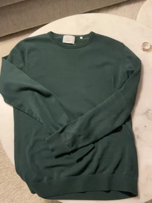 Mörkgrön stickad tröja från Bläck - Mörkgrön, stilren stickad tröja från Bläck med rund halsringning och långa ärmar. Tröjan är tillverkad i ett mjukt, tunt material och har ribbade muddar vid hals, ärmslut och nederkant. Perfekt för en enkel och clean look. Nypris 599kr! Knappt använd