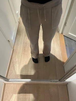 Beige juicy byxor med fickor - Säljer ett par beige mjukisbyxor med dragsko i midjan och stora fickor fram. Byxorna har även en bakficka med knapp. Sköna och avslappnade med rak passform, perfekta för chill dagar. Materialet känns mjukt och bekvämt.