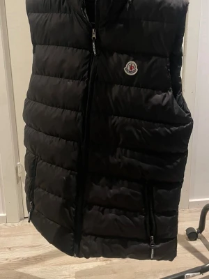 Svart dunväst från Moncler - Snygg svart dunväst från Moncler med klassisk logga på bröstet. Västen har hög krage, dragkedja framtill och två fickor med dragkedja. Perfekt för lager-på-lager och riktigt skön att bära under kyliga dagar.