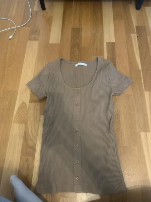 Beige ribbad topp med knappar - Snygg beige ribbad topp med korta ärmar och rund halsringning. Toppen har knappar framtill hela vägen ner och är i ett stretchigt material som sitter skönt på kroppen. 