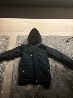 Svart jacka med huva från Dope - Snygg svart jacka från Dope med huva och flera praktiska fickor framtill. Jackan har dragkedja och tryckknappar, samt logga på bröstet och ärmen. Perfekt för kalla dagar och outdoor-aktiviteter. Materialet är slitstarkt och vattenavvisande.