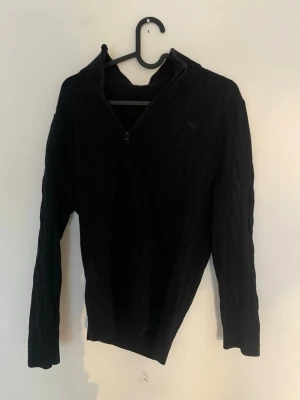 Svart kabelstickad tröja med halvzip - Skriv om ni vill ha mått Svart kabelstickad tröja med halv dragkedja och hög krage. Tröjan har långa ärmar och ribbstickade muddar vid ärmslut och nederkant. Perfekt för lager-på-lager och en stilren look.