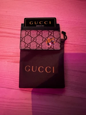 Gucci plånbok med tigerdetalj - Säljer en snygg Gucci plånbok i beige och brunt med klassiskt GG-mönster och en cool tigerdetalj på framsidan. Plånboken är i canvasmaterial och har plats för kort. Kommer med originalpåse från Gucci.
