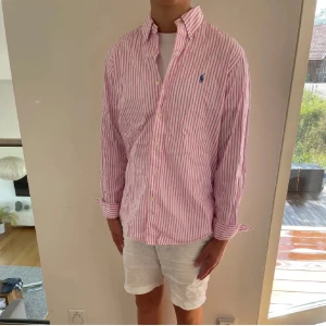 Rosa och vit randig skjorta från Polo Ralph Lauren - Snygg skjorta med rosa och vita vertikala ränder från Polo Ralph Lauren. Klassisk krage, långärmad och med knappar framtill. Broderad blå logga på bröstet. Skjortan är i mjuk bomull och har en normal passform som funkar till många stilar. Obs croppad i längden, exakt längd på skjortan kommer inom kort 