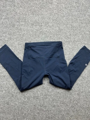 Mörkblå leggings från Lululemon - Mörkblå leggings från Lululemon med hög midja och smickrande sömmar. Tillverkade i ett stretchigt funktionsmaterial som andas. Snygg passform och praktiska sidofickor. Liten Lululemon-logga på benet.