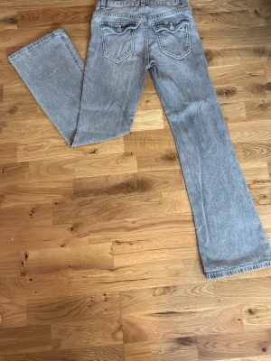 Grå bootcut jeans med fickdetaljer - Säljer ett par grå bootcut jeans med snygga fickdetaljer bak och klassisk femficksmodell. Jeansen har en ljus tvätt och raka ben med lätt utsvängning. Perfekta för en avslappnad och trendig look.