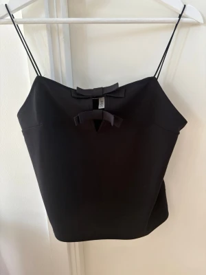 Svart linnetopp med rosetter - Snygg svart linnetopp med tunna axelband och två dekorativa rosetter framtill. Toppen har en enkel och stilren design som passar perfekt till många olika outfits. Toppen är ny utan prislapp🖤