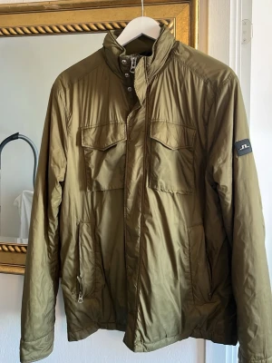 Olivgrön jacka från J.Lindeberg - Snygg olivgrön vindjacka från J.Lindeberg med hög krage och två bröstfickor med lock och knapp. Jackan har en stilren design med diskret logotyp på ärmen och är tillverkad i ett lätt, glansigt material som skyddar mot vind. Några små märken/litet hål. (Se bild) annars bra skick hålet i bröst fickan på knappen syns inte för den är under fickan på knappen, sen det lilla rivmärket är inte heller så synligt då du har armen där naturligt. 