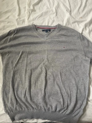 Grå stickad tröja Tommy Hilfiger - Snygg grå stickad tröja från Tommy Hilfiger med V-ringning och diskret logga broderad på bröstet. Tröjan har ribbade muddar vid ärmslut och nederkant samt klassisk passform. Perfekt till lager på lager och enkel att matcha.