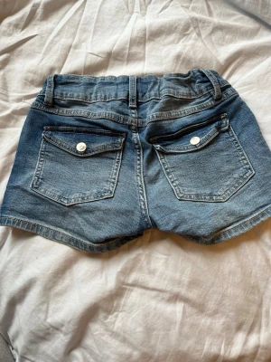 Blå denimshorts med dubbla knappar - Snygga blå jeansshorts i klassisk denim med dubbla knappar framtill och fem fickor. Shortsen har normal passform, samt ficklock med knapp bak. Perfekta för sommaren och lätta att matcha. Det är justerbara i midjan