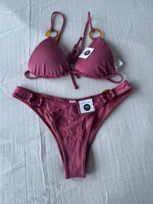 Rosa bikini med ringdetaljer XS - Trendig rosa bikini från Bik Bok i storlek XS. Bikinin har trekantsöverdel med knyt och snygga ringdetaljer i plast både på överdel och underdel. Perfekt för strandhäng och poolparty. Materialet känns mjukt och följsamt mot huden.