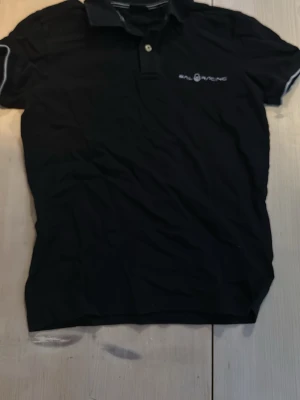Svart pikétröja från Sail Racing - Klassisk svart pikétröja från Sail Racing med vit logga på bröstet och vita detaljer på ärmarna. Tröjan har krage, korta ärmar och knäppning framtill. Perfekt till jeans eller shorts för en clean look.