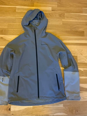 Grå vind/regnjacka från Peak Performance - Snygg grå vindjacka från Peak Performance med svart dragkedja och två sidofickor med dragkedja. Jackan har huva och ljusare grå partier på ärmarna. Perfekt för blåsiga dagar och outdoor-aktiviteter.