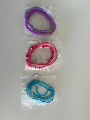 3 förpackningar (8st) Färgglada armband - Tre armband i olika färger: lila, rosa/vit och turkos. Alla armband är gjorda av platta pärlor i plast och har en modern, ungdomlig stil. Perfekta för att ge din outfit en färgklick och personlig touch.