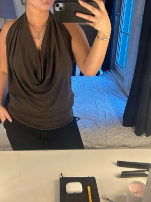 Brun draperad ärmlös topp - Säljer en brun ärmlös topp med snygg draperad front och halterneck. Toppen har en avslappnad passform och är tillverkad i ett mjukt, stretchigt material som känns skönt mot huden. Perfekt att styla med jeans eller kjol för en trendig look. Nypris 329 från Gina, prislappen sitter kvar. 