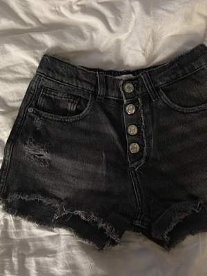 Svarta högmidjade jeansshorts - Säljer ett par svarta högmidjade jeansshorts med fransiga kanter och slitningar. Shortsen har fyra silverfärgade knappar framtill och klassiska fem fickor. Perfekta för sommaren och ger en cool, avslappnad look.