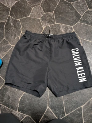 Svarta badbyxor Calvin Klein stl 14-16 år - Snygga svarta badbyxor från Calvin Klein med vit logga längs ena benet. Klassisk modell med resår och dragsko i midjan samt meshfoder på insidan. Perfekta för stranden eller poolen, tillverkade i snabbtorkande material. nya endast provade 