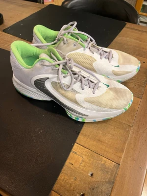 Nike sneakers i vitt och grönt - Snygga Nike basketskor med vit mesh och ljusgrå detaljer, grönt innerfoder och coola gröna och blå inslag på sulan. Skorna har en sportig design med låg profil och stora svarta swoosh-loggor på sidorna. Perfekta för dig som gillar streetstyle och sportig look.