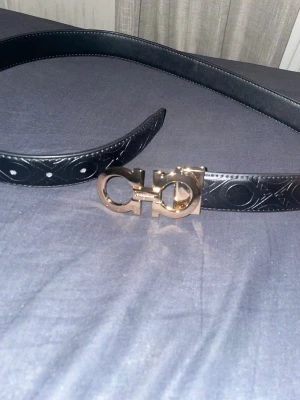 Svart bälte med guldfärgat spänne - (105cm) Snyggt svart skärp med präglat mönster längs hela remmen och ett ikoniskt, guldfärgat spänne. Skärpet är gjort i skinn och har en elegant och modern look som lyfter vilken outfit som helst. 