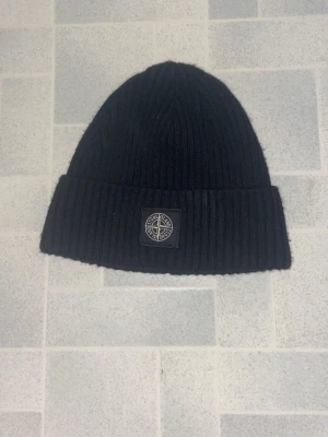 Svart ribbstickad mössa Stone Island - Svart ribbstickad mössa från Stone Island med uppvikt kant och klassisk logopatch framtill. Snygg och enkel design som passar till det mesta. Perfekt för kalla dagar när du vill hålla stilen.