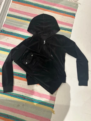 Svart set från Juicy Couture - Mysigt svart set i velour från Juicy Couture. Setet består av en hoodie med den klassiska dragkedjan, huva och fickor samt matchande byxor med fickor med knappar. 