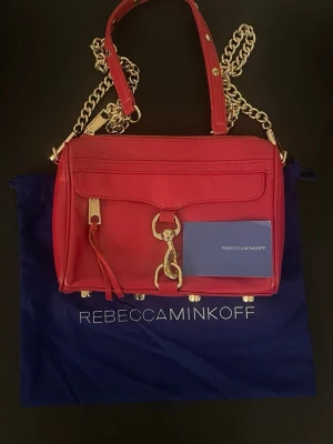 Röd handväska från Rebecca Minkoff - Cool röd handväska i läder från Rebecca Minkoff med guldfärgade kedjor och detaljer. Väskan har en dragkedja upptill, dekorativt spänne framtill och en ficka på framsidan. Perfekt storlek för det viktigaste och snygg att bära crossbody eller i handen.