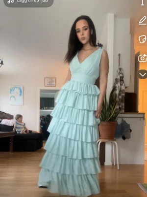  balklänning med volanger - Superfin långklänning i mintgrönt med djup v-ringning och markerad midja. Klänningen har flera lager volanger som ger en riktigt drömmig och flowy känsla. Perfekt för dig som vill sticka ut med en romantisk och elegant look.