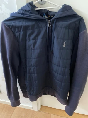 Mörkblå cardiganjacka Polo Ralph Lauren - Mörkblå cardiganjacka från Polo Ralph Lauren med quiltad front och dragkedja. Jackan har huva med dragsko, mjuka ärmar i sweatshirtmaterial och broderad logga på bröstet. Perfekt för en stilren och avslappnad look.