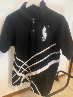 Svart pikétröja från Ralph Lauren - Svart pikétröja från Ralph Lauren med vit broderad logga på bröstet och vita grafiska linjer framtill. Tröjan har krage, knappar och korta ärmar samt en stor vit siffra på ena ärmen. Klassisk och sportig stil.