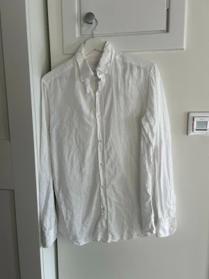 Vit linneskjorta från Massimo Dutti - Klassisk vit skjorta från Massimo Dutti i 100% linne. Skjortan har lång ärm, knappar framtill och en stilren krage. Perfekt för en fräsch och avslappnad look under varmare dagar.