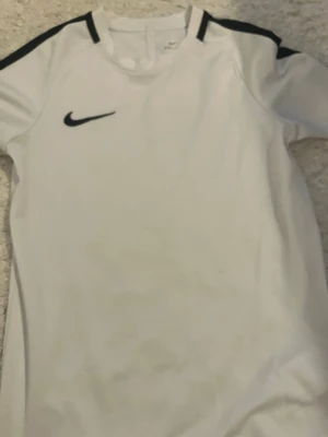 Vit Nike Dri-Fit sporttröja - Säljer en vit sporttröja från Nike med svarta detaljer på axlarna och svart Nike-logga på bröstet. Tillverkad i Dri-Fit-material som andas och passar perfekt till träning eller match. Tröjan har rund hals och korta ärmar.