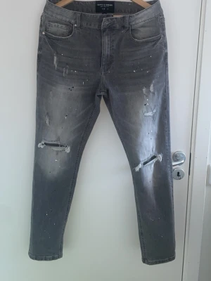 Grå jeans från  Supply & Demand - Säljer ett par grå jeans från Supply & Demand med slitningar och hål på knäna samt coola färgstänk. Jeansen har fem fickor och en klassisk passform. Perfekta för dig som gillar en trendig och avslappnad stil.