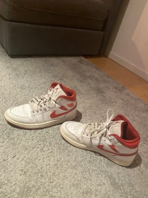 Nike Air Jordan 1 Mid Vit/Röd - Säljer ett par Nike Air Jordan 1 Mid sneakers i vitt läder med röda detaljer och swoosh. Skorna har perforerad tå, röda innersulor och yttersula samt klassisk Jordan-logga på sidan. Perfekta för dig som gillar streetwear och snygg retrostil.