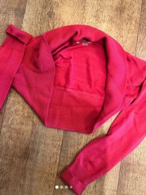 Rosa bolero med bred krage - Kort bolero i starkt rosa färg med bred ribbad krage och muddar. Långärmad modell som passar perfekt över linnen eller klänningar. Materialet ser ut att vara mjukt och stretchigt stickat tyg.